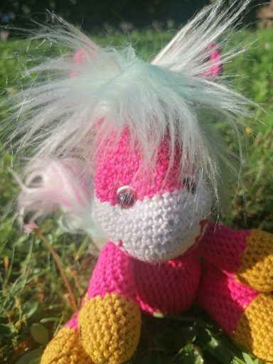 Häkelpferd mit buntem Körper und buntem, flauschigem Haar in einer grünen Umgebung.