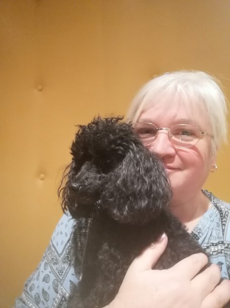 Frau mit blonden Haaren umarmt einen schwarzen Pudel vor einem gelben Hintergrund.