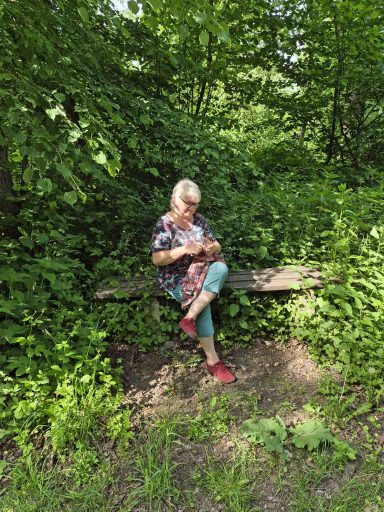 Frau sitzt auf einer Bank in einem dichten, grünen Wald mit Pflanzen um sie herum.