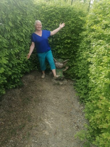 Person mit blauer Bluse und grünen Shorts steht in einem grünen Gartenlabyrinth.