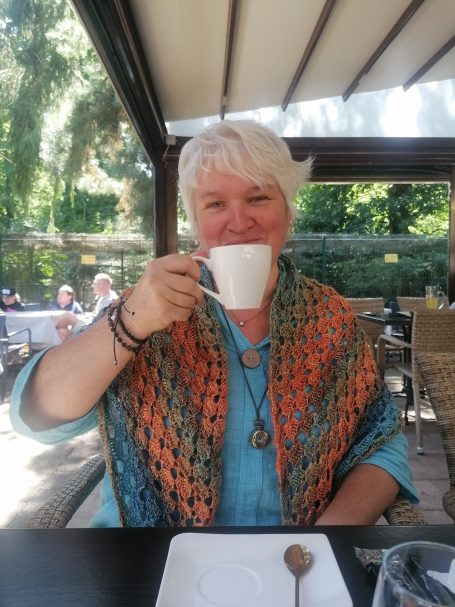 un ä Gäffchen Frau mit weißem Haar, hält eine Tasse Kaffee und lächelt, in einem Gartenrestaurant.